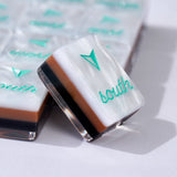 A close-up of a square acrylic keycap with a white top, a brown middle band, a black bottom band, and a teal logo and the word 'south' on a white surface.
#color_tortoiseshell pearl edition