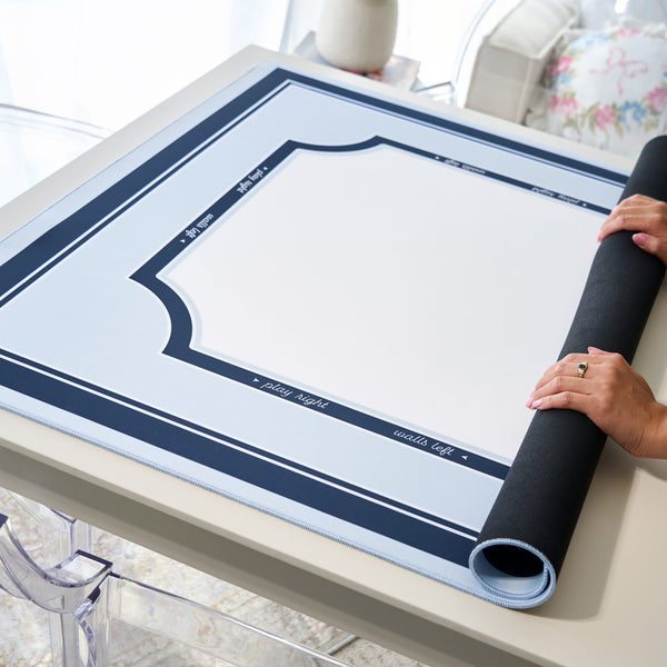 A large white and navy blue photo mat with decorative border lines is laid out on a table, with a person's hands rolling up one of the black protective sleeves on the right side.
#color_classic carolina blue