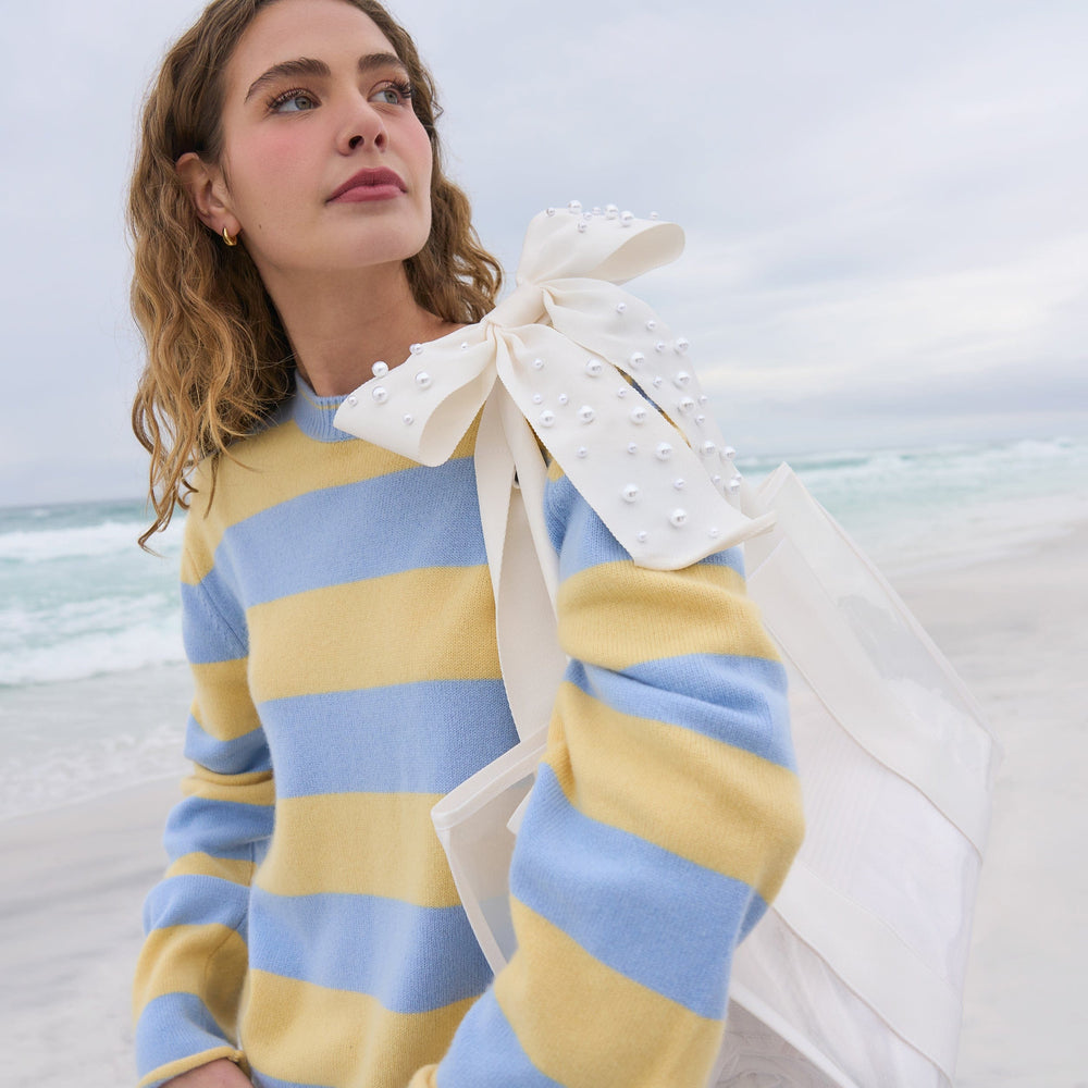 Woman in a blue and yellow striped dress with a large tote with a white pearly bow on a beach.