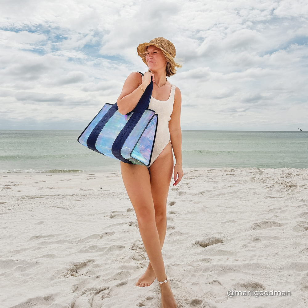 Woman standing in the sand on a sunny day at the beach with a jelly tote on her shoulder and a straw hat on 
#color_oxford navy iridescent