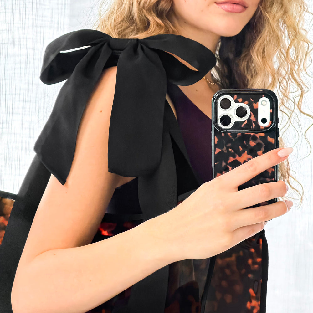Woman holding a phone with a black case featuring a Tote with bow detail on her shoulder. 

#color_tortoiseshell bow