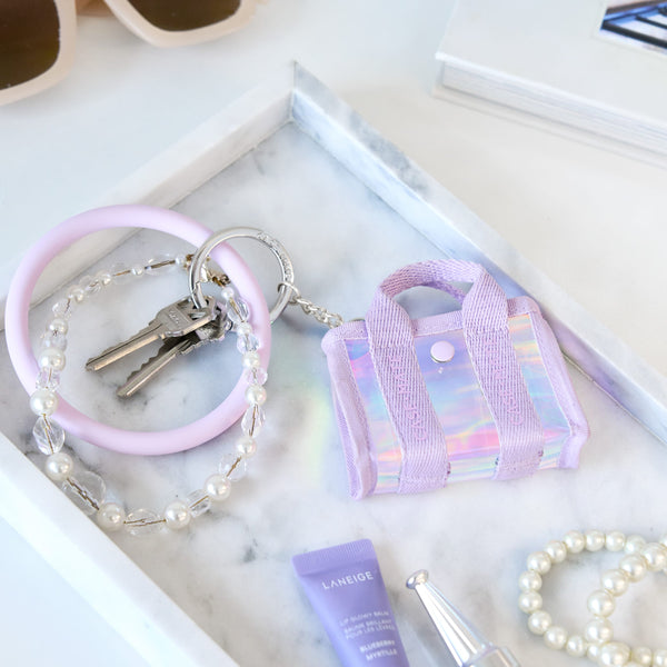 Keychain with pink bag charm, keys, and bracelet on a marble surface with a bottle and book.
#color_iridescent