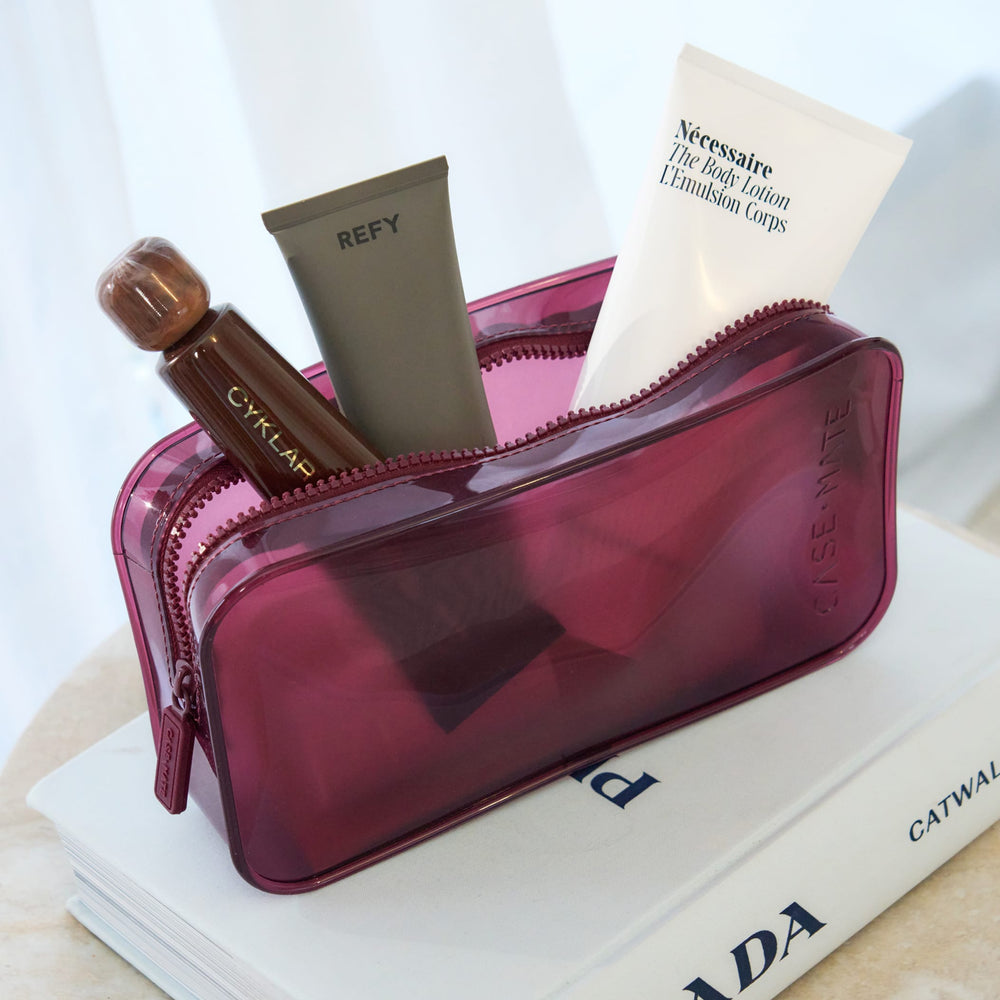 A translucent magenta cosmetic pouch filled with skincare items, including a white tube labeled Nécessaire and a brown bottle labeled CYKLAR, styled on top of a white book.

#color_mulberry