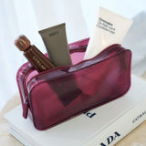 A translucent magenta cosmetic pouch filled with skincare items, including a white tube labeled Nécessaire and a brown bottle labeled CYKLAR, styled on top of a white book.

#color_mulberry