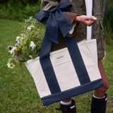 A person outdoors carrying a large off-white tote bag with bold navy blue side panels and handles, tied with a matching navy bow. The tote has a front pocket with the visible brand name 'CASE-MATE' and is filled with wildflowers.
#color_oxford navy bow