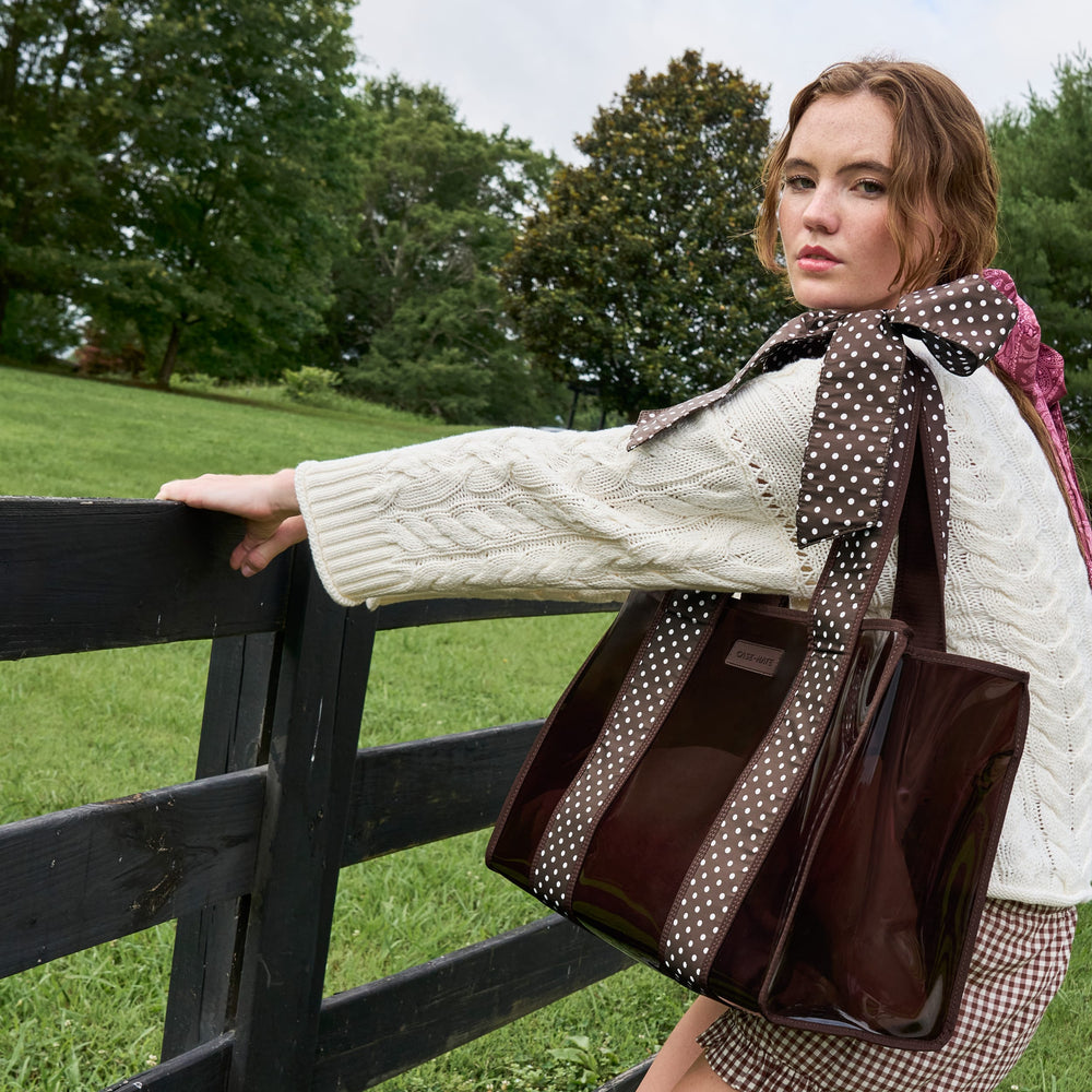 A woman outdoors by a wooden fence wearing a cream cable-knit sweater and holding a glossy dark brown tote bag with brown-and-white polka-dot shoulder straps.
#color_espresso polka dot bow