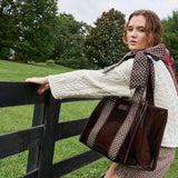 A woman outdoors by a wooden fence wearing a cream cable-knit sweater and holding a glossy dark brown tote bag with brown-and-white polka-dot shoulder straps.
#color_espresso polka dot bow