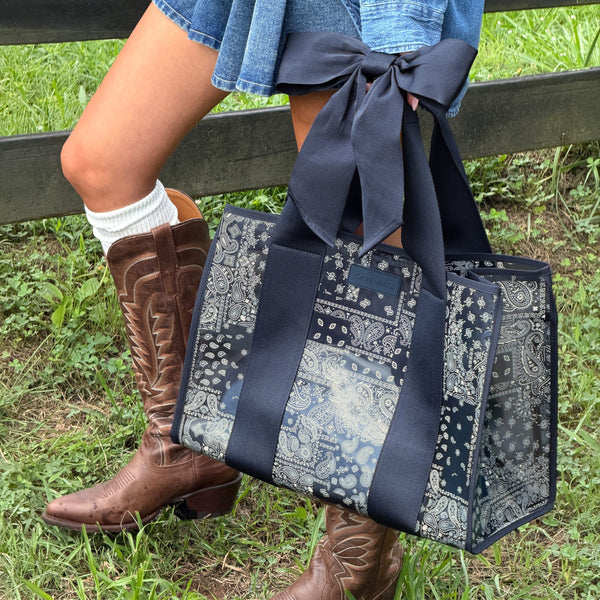 A navy blue tote bag with a bandana print pattern and large bow handle, held by a person wearing brown cowboy boots and a denim skirt in an outdoor grassy setting.
#color_navy bandana bow