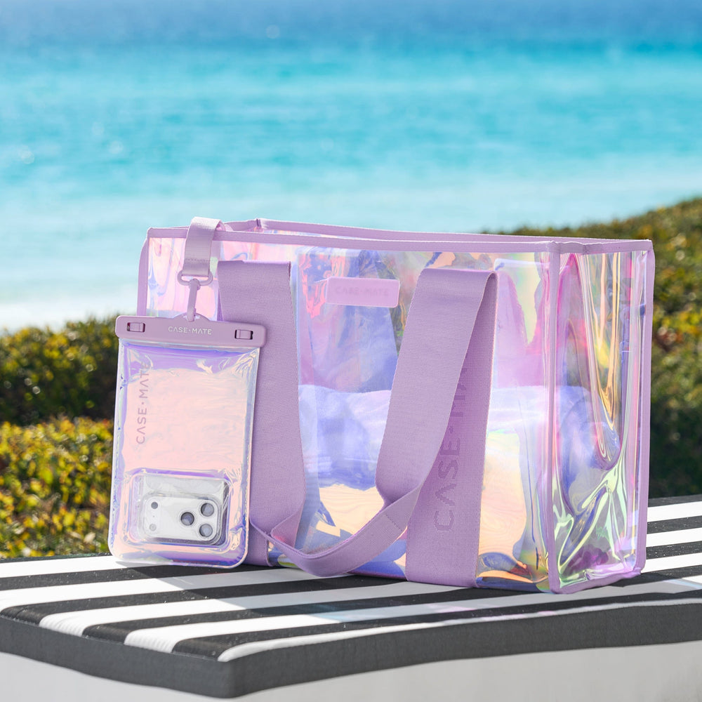 Clear holographic beach bag with a phone case on a striped surface with a blue sky background
