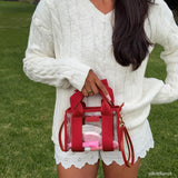 A person outdoors on grass holding a small transparent tote bag with bright red handles, straps, and trim. The person is wearing a white cable-knit sweater and white eyelet shorts.

#color_varsity red bow