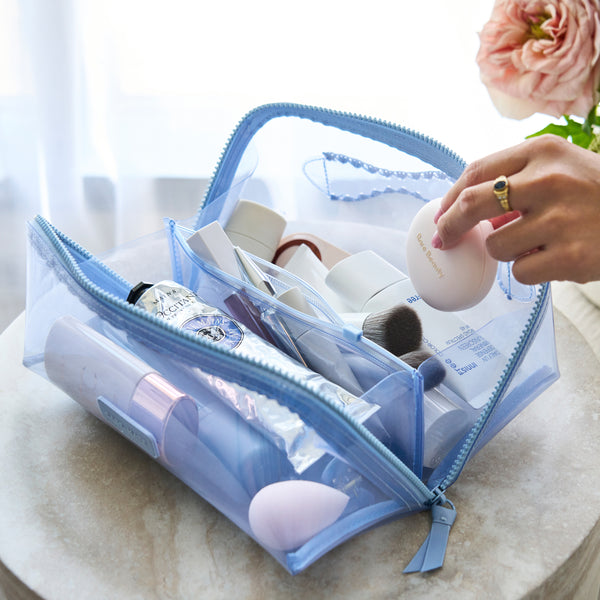 A light blue translucent zippered makeup bag filled with cosmetics, shown open on a marble surface with a hand reaching toward a makeup sponge inside.
#color_carolina blue crochet wave