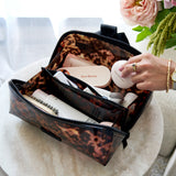 A leopard-print cosmetic bag filled with beauty products, shown on a round table with a hand placing a white round item inside. The background includes soft-focus flowers and a light-colored room.
#color_tortoiseshell 