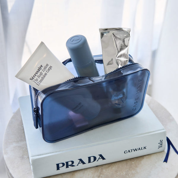 A translucent navy blue cosmetic pouch filled with toiletries, and a gray bottle, resting on top of a white Prada book.

#color_oxford navy
