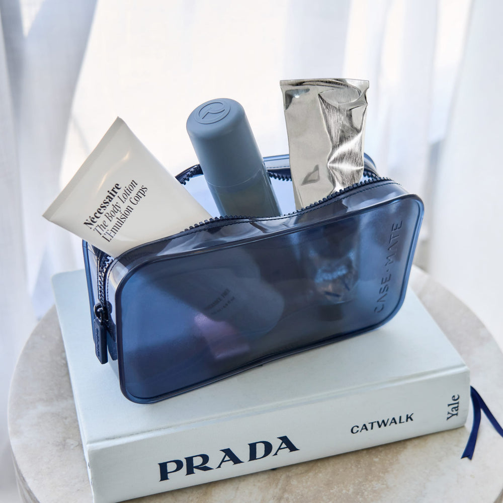 A translucent navy blue cosmetic pouch filled with toiletries, and a gray bottle, resting on top of a white Prada book.

#color_oxford navy