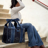 A woman sits on outdoor stone stairs wearing a white long-sleeve top and blue jeans, with a large navy tote bag featuring a prominent bow handle resting beside her.
#color_dark wash denim bow