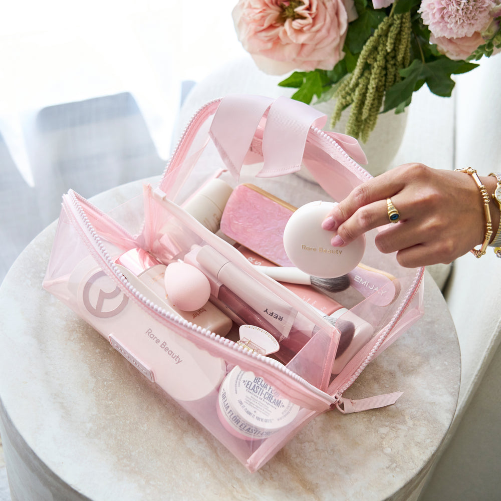 A pink translucent makeup bag filled with cosmetics, placed on a round marble table. A person's hand is holding one of the cosmetic items inside the bag.
#color_ballet pink bow