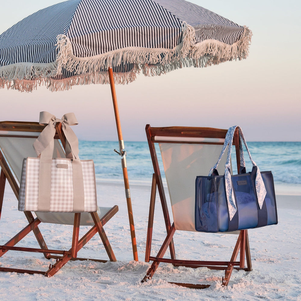 Two totes hanging on two beach chairs on the beach with an umbrella between the chairs