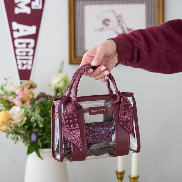 A hand holding a maroon and clear tote bag with matching maroon bandana handles and a lace insert, set against a softly blurred indoor background with flowers and a pennant.
#color_mulberry twilly