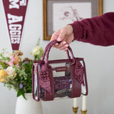 A hand holding a maroon and clear tote bag with matching maroon bandana handles and a lace insert, set against a softly blurred indoor background with flowers and a pennant.
#color_mulberry twilly