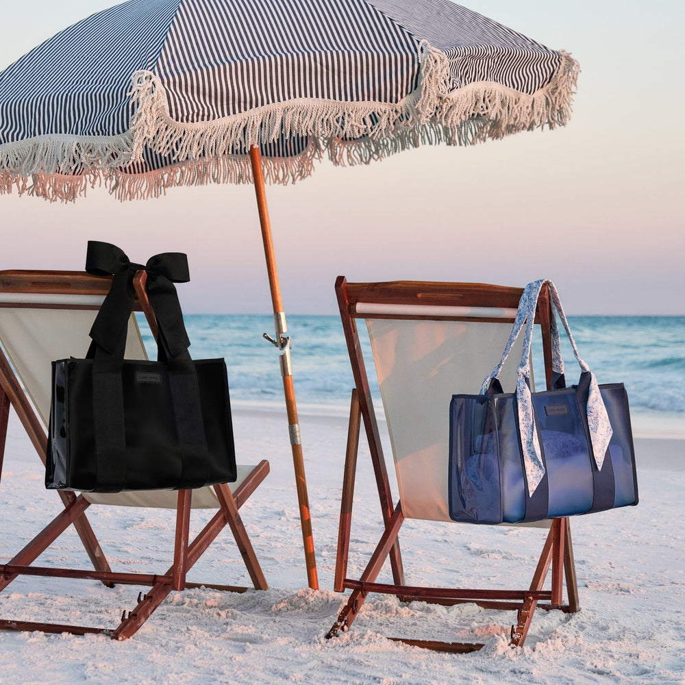 Two beach chairs with black and blue bags under a striped umbrella on a sandy beach.