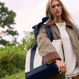 A woman outdoors carrying a large cream and navy tote bag over her shoulder, with a white sweater and olive jacket visible. The background shows blurred greenery and sky.
#color_oxford navy bow