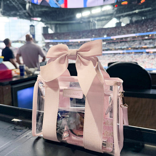 A pink and clear tote bag with a large pink bow on a stadium table during a game, featuring a visible Rose Nate brand label.
#color_ballet pink bow