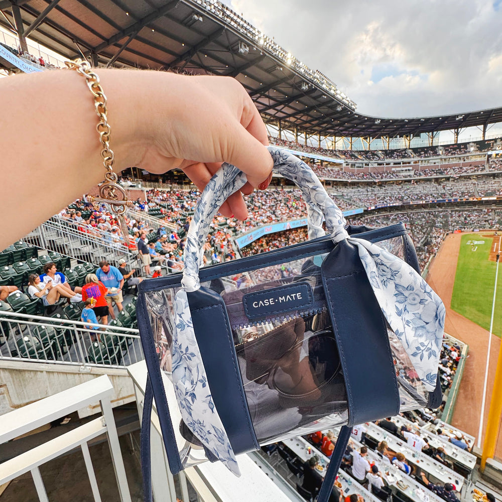 A hand holds a small transparent handbag with navy blue trim and a floral fabric handle in a crowded outdoor stadium. The bag has a visible CASE MATE logo plaque on the front.
#color_something navy twilly