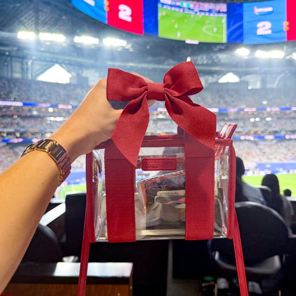 A clear tote bag with a large red ribbon bow and red straps, held by a person's hand in a stadium setting. The bag has a visible 'Society6' logo on the front pocket.

#color_varsity red bow