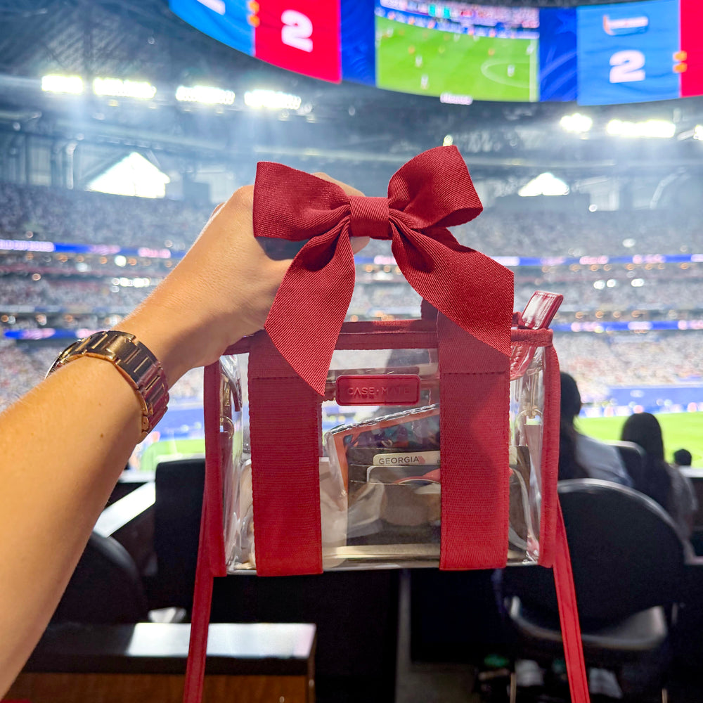 A clear tote bag with a large red ribbon bow and red straps, held by a person's hand in a stadium setting. The bag has a visible 'Society6' logo on the front pocket.

#color_varsity red bow
