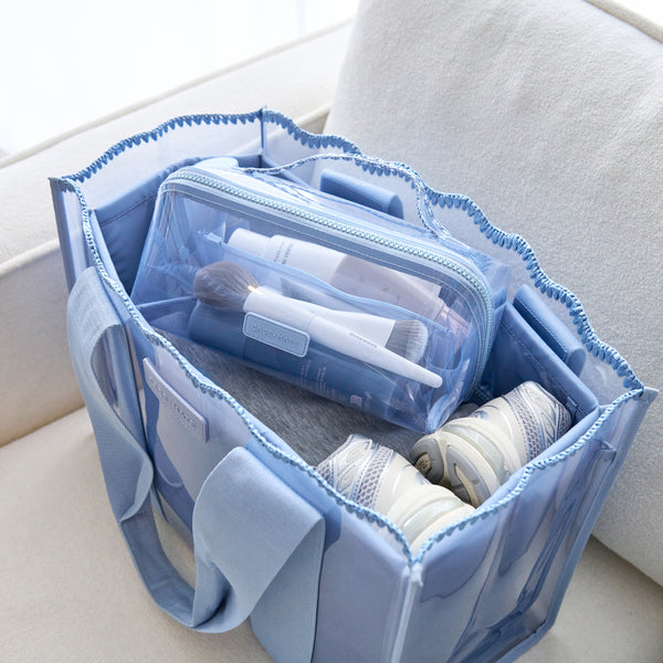 A light blue fabric toiletry bag with clear zippered pockets, shown open and filled with travel-sized toiletries on a light beige sofa.
#color_carolina blue crochet wave