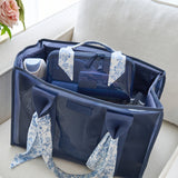 A navy blue diaper bag with clear plastic side panels and floral fabric handles, shown open on a light beige sofa with baby accessories inside.
#color_something navy twilly