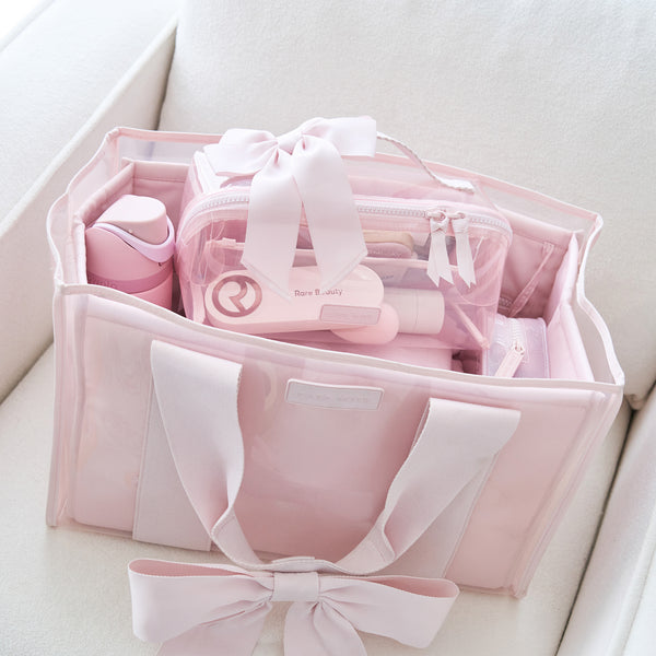 A light pink tote bag with satin ribbon handles and a matching clear zippered pouch filled with pink toiletries, placed on a white upholstered chair.
#color_ballet pink bow