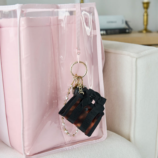 A pink translucent zippered tote bag with a black and brown tortoiseshell miniature suitcase keychain charm attached to the key leash, resting on a light beige sofa

#color_tortoiseshell bow