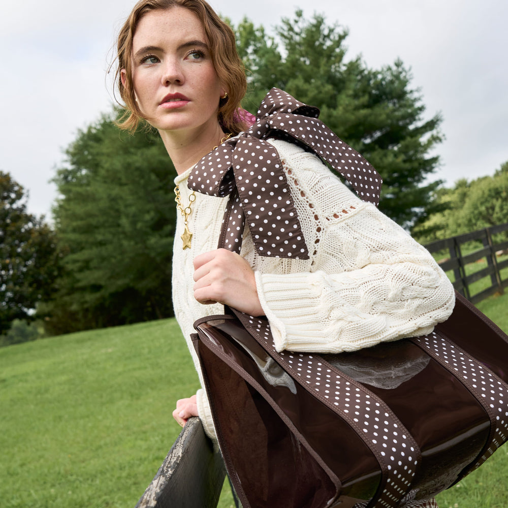 A woman outdoors wearing a cream cable-knit sweater and carrying a brown and white polka-dot jelly tote bag over her shoulder. 
#color_espresso polka dot bow