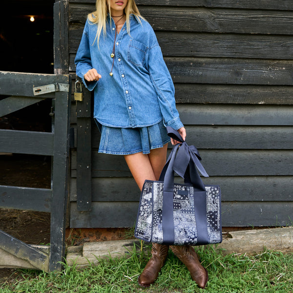 A woman wearing a denim shirt and denim skirt stands in front of a dark wooden barn door, holding a large navy and white patterned tote bag with bow handles.
#color_navy bandana bow