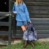 A woman wearing a denim shirt and denim skirt stands in front of a dark wooden barn door, holding a large navy and white patterned tote bag with bow handles.
#color_navy bandana bow