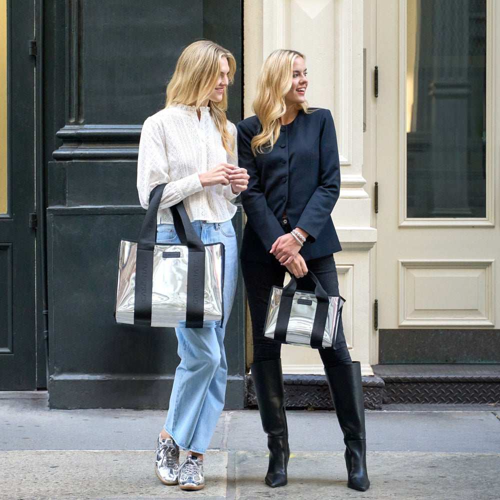 Two women holding silver handbags with black straps in front of a building.