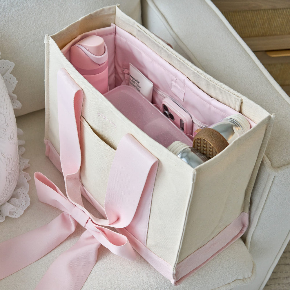 A pink and cream canvas tote bag with an organizer inside, shown on a light upholstered chair. The bag is open and filled with pink accessories, including a bottle, pouches, and small items.