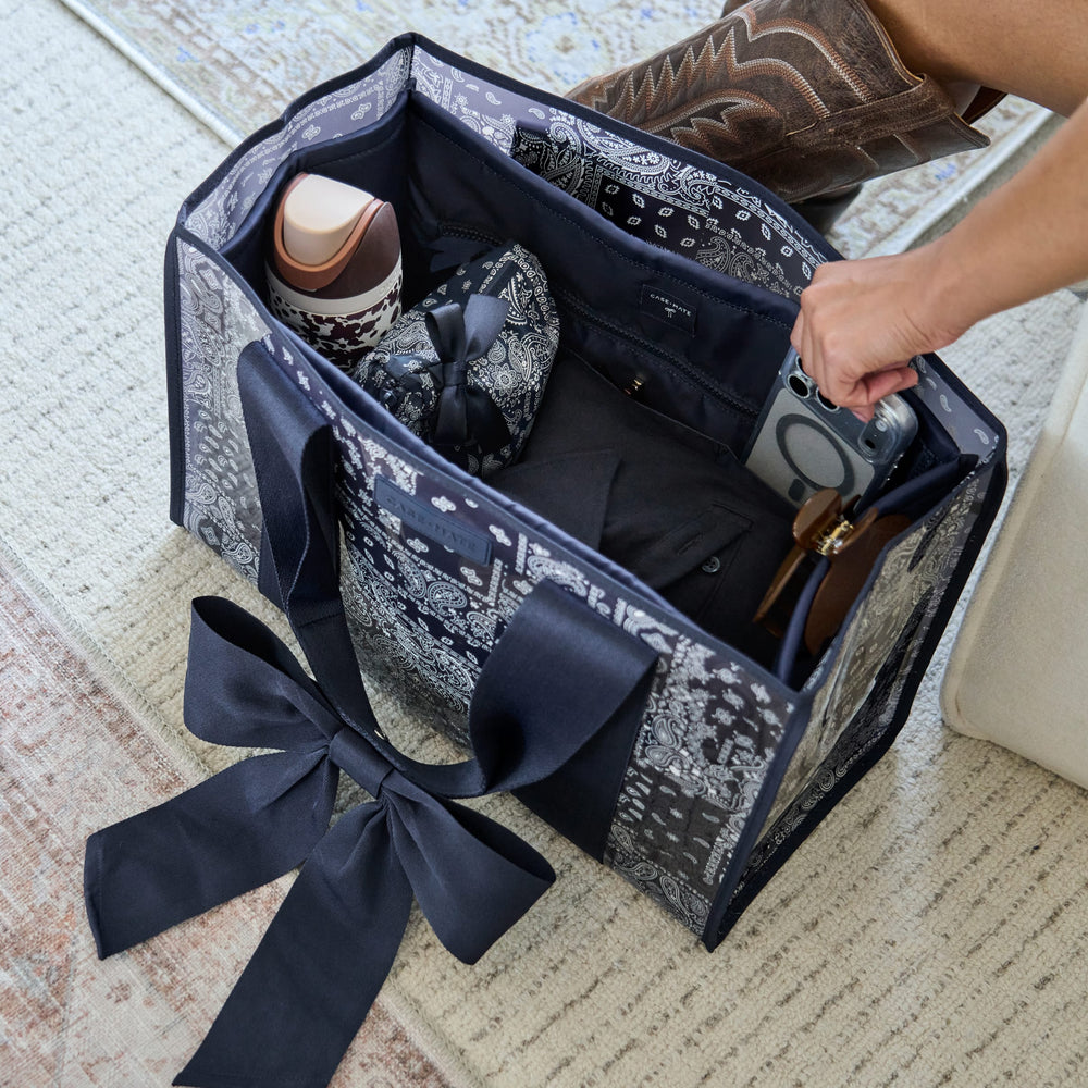 A navy and white patterned paisley travel tote bag with a large navy bow, shown open on a textured rug with a person's hand reaching inside and a brown cowboy boot visible at the top right.