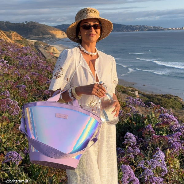 Woman standing in a scenic coastal area with a colorful bag and drink, wearing a hat.
#color_iridescent