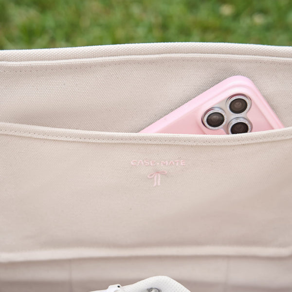 Close-up of a beige canvas shoulder bag with a pink smartphone tucked into the side pocket. #color_ballet pink bow