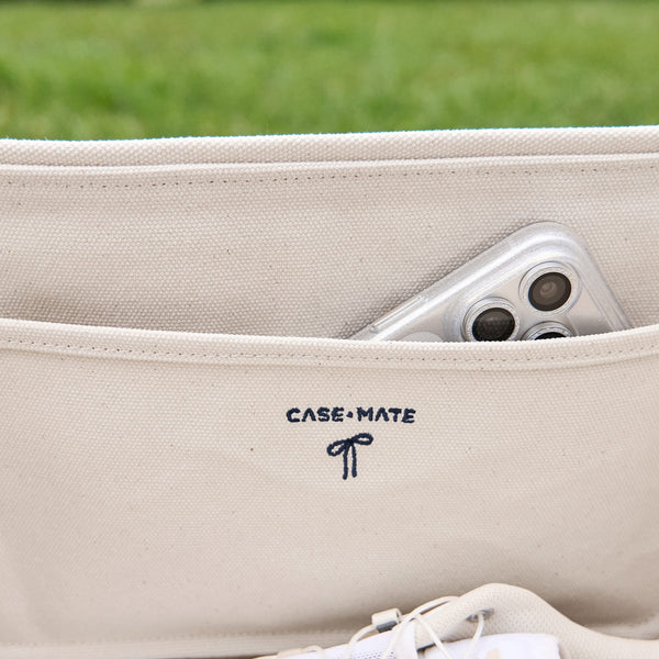 Close-up of a beige canvas laptop sleeve with the CASE-MATE logo embroidered in navy on the front, showing a silver smartphone tucked into a side pocket, with a blurred green grass background.
#color_oxford navy bow