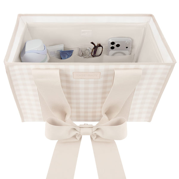 A square fabric storage organizer bin with a beige and white gingham pattern, featuring wide cream-colored shoulder straps tied into a large bow at the front. The open top of the bin is shown holding various small personal items like a phone, glasses, and a small bottle.

#color_french vanilla 