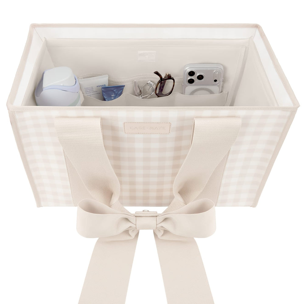 A square fabric storage organizer bin with a beige and white gingham pattern, featuring wide cream-colored shoulder straps tied into a large bow at the front. The open top of the bin is shown holding various small personal items like a phone, glasses, and a small bottle.

#color_french vanilla 