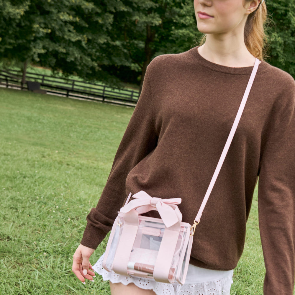 A woman wearing a brown knit sweater and white skirt carries a small transparent pink crossbody bag with a bow on a strap in an outdoor park setting.
#color_ballet pink bow