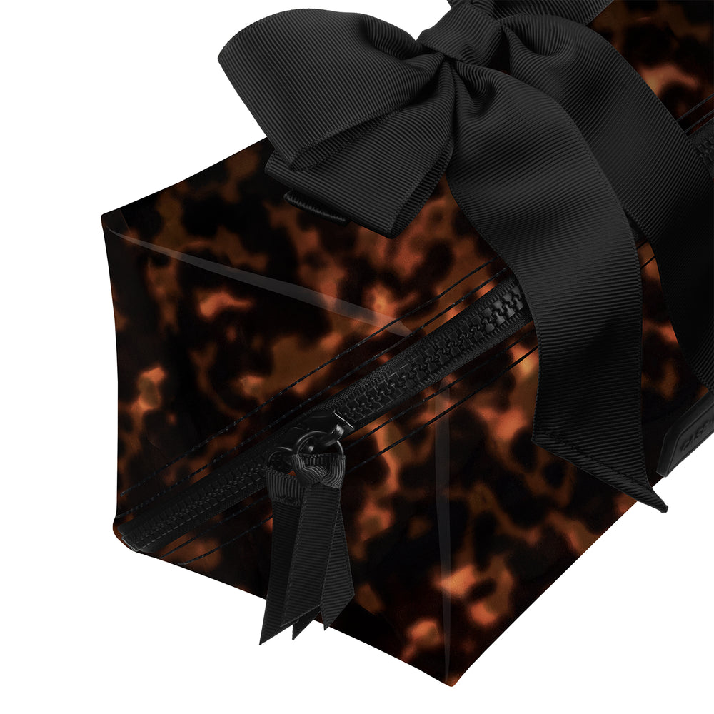 A clear acrylic zippered pouch with a black and orange tortoiseshell pattern, decorated with a large black satin bow and a black ribbon zipper pull, shown on a white background.
#color_tortoiseshell 