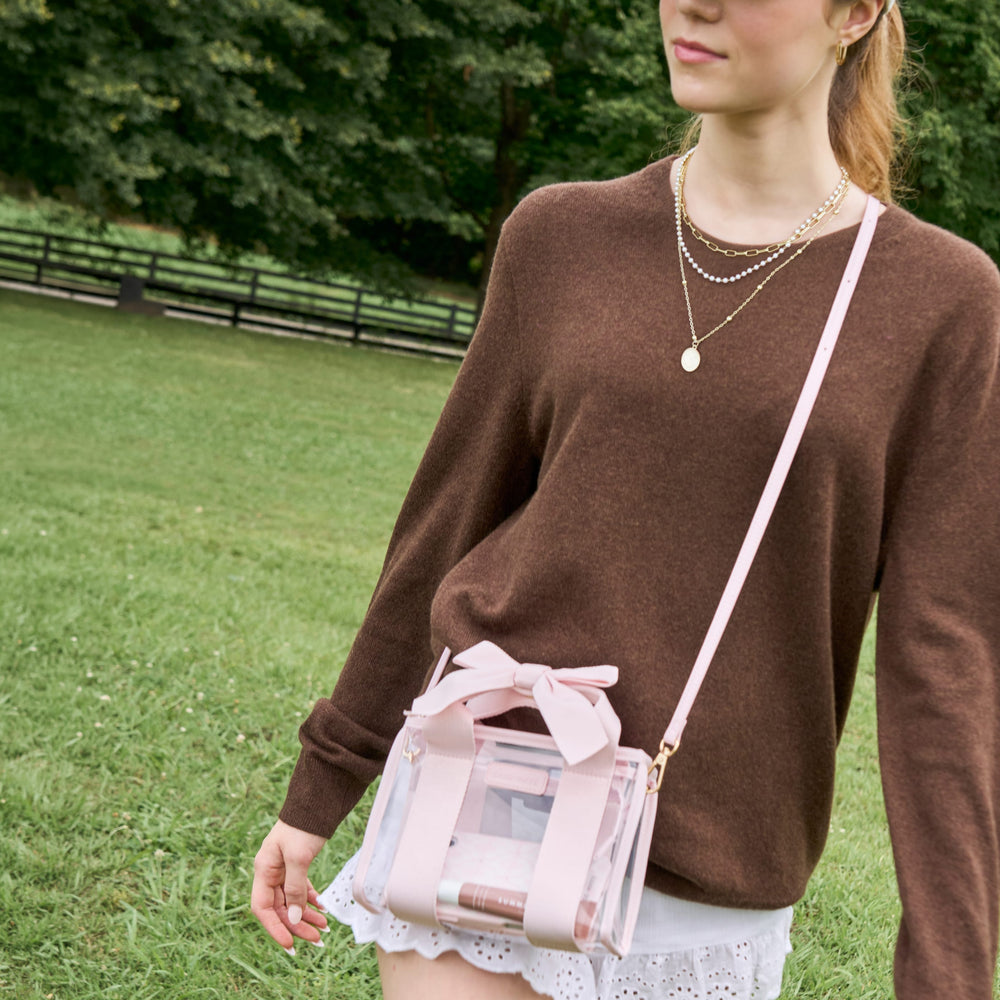 A woman wearing a brown knit sweater and white skirt carries a small transparent pink crossbody bag with a bow on a strap in an outdoor park setting.
#color_ballet pink bow