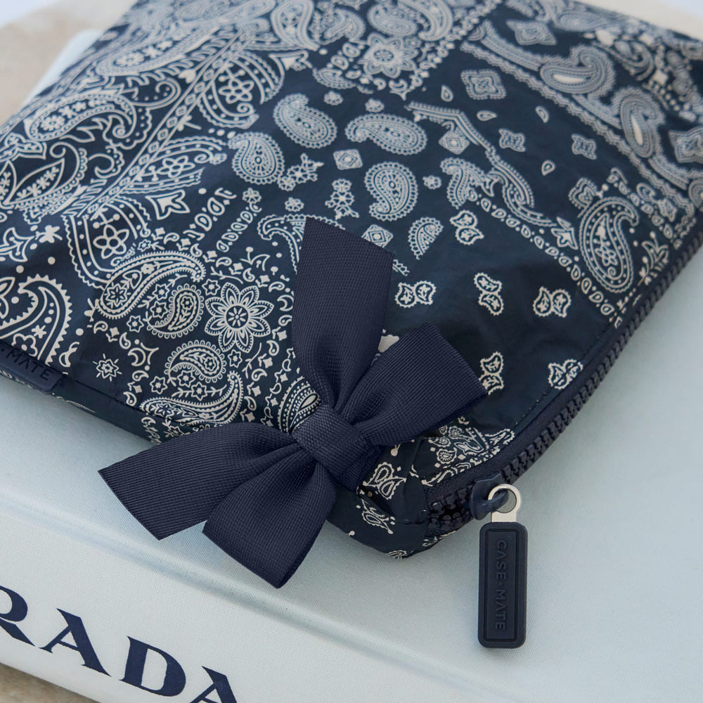 A navy blue and white paisley bandana-patterned zippered pouch with a matching fabric bow and a black zipper pull tag, resting on a white book. 
#color_navy bandana bow