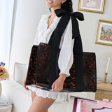 A woman in a white ruffled blouse and skirt carries a large tote bag with black shoulder straps and tortoiseshell-patterned sides, photographed in a bright indoor room.
#color_tortoiseshell bow