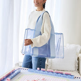 A woman wearing a cream knit sweater and blue jeans carries a large, clear plastic tote bag with light blue straps in a bright, modern room.

#color_carolina blue crochet wave